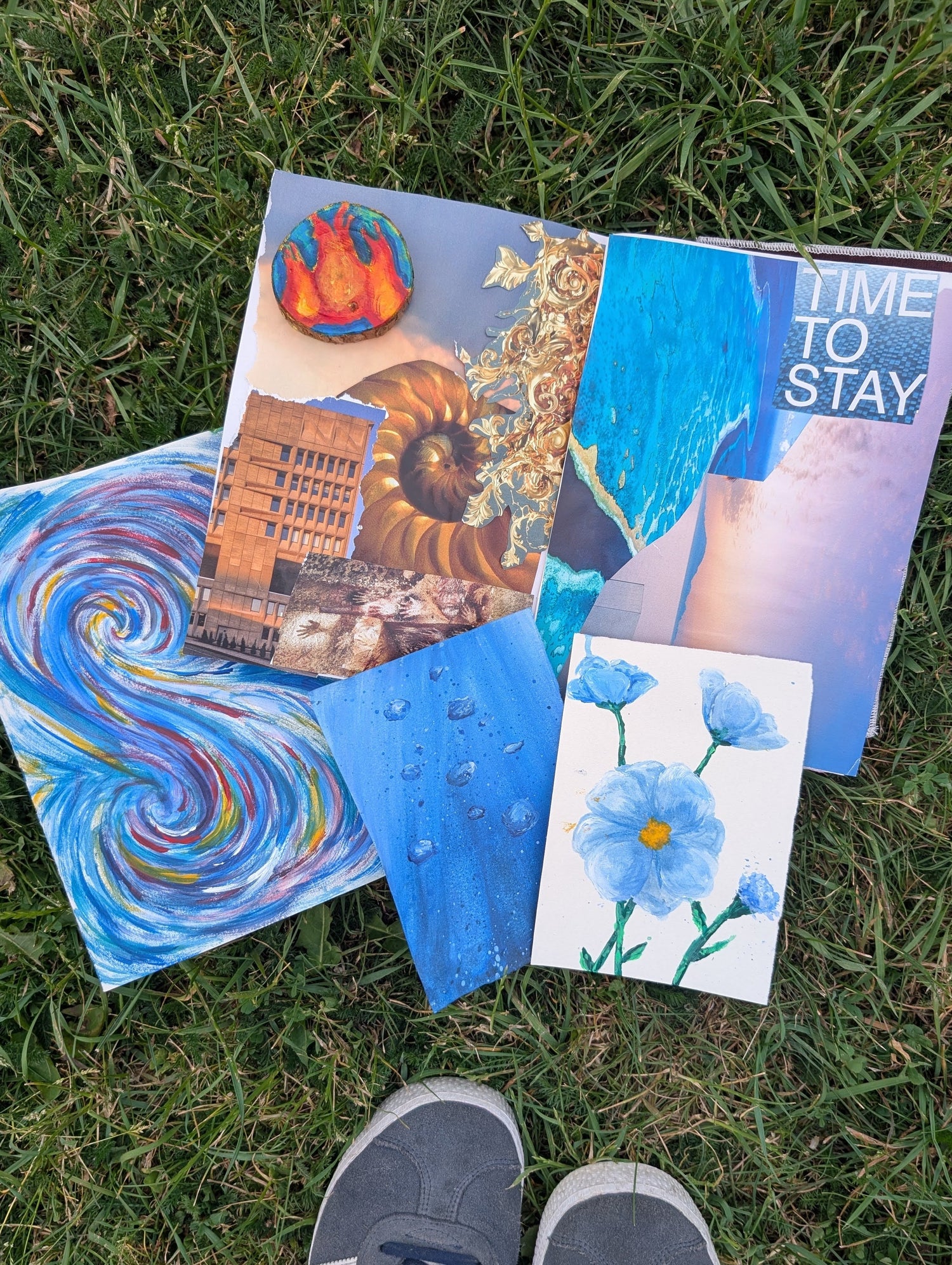 Several colorful art prints and paintings are arranged on green grass, with a person's gray sneakers visible at the bottom of the frame. The artwork includes abstract blue swirls, floral blue-and-white flowers, a shell and cityscape collage, and a blue-and-white text print reading 'TIME TO STAY'.