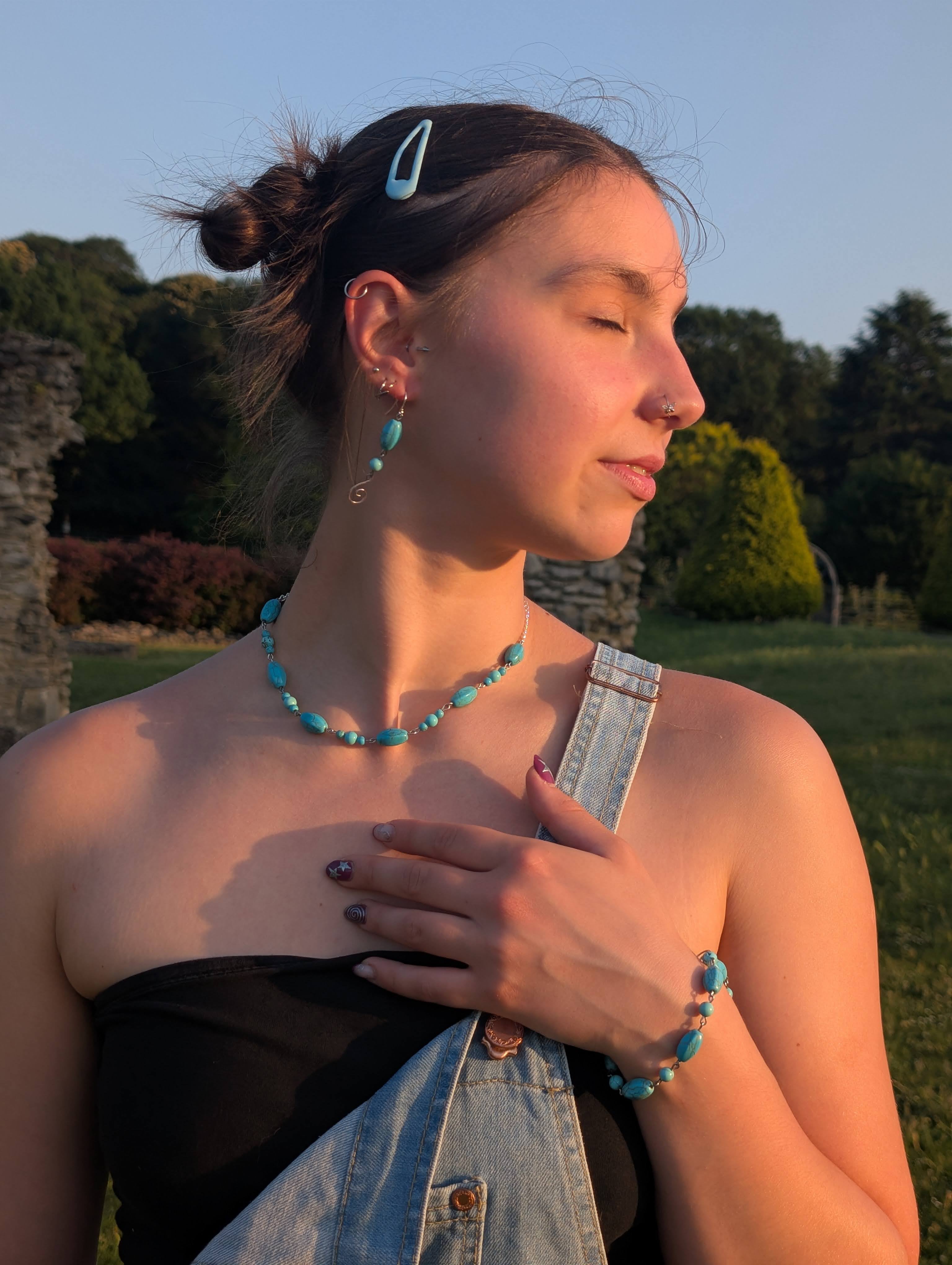 A woman outdoors wearing a matching turquoise beaded necklace, bracelet, and earring, with a turquoise hair clip. She is wearing a black strapless top and denim overalls, with trees and a stone wall in the background.